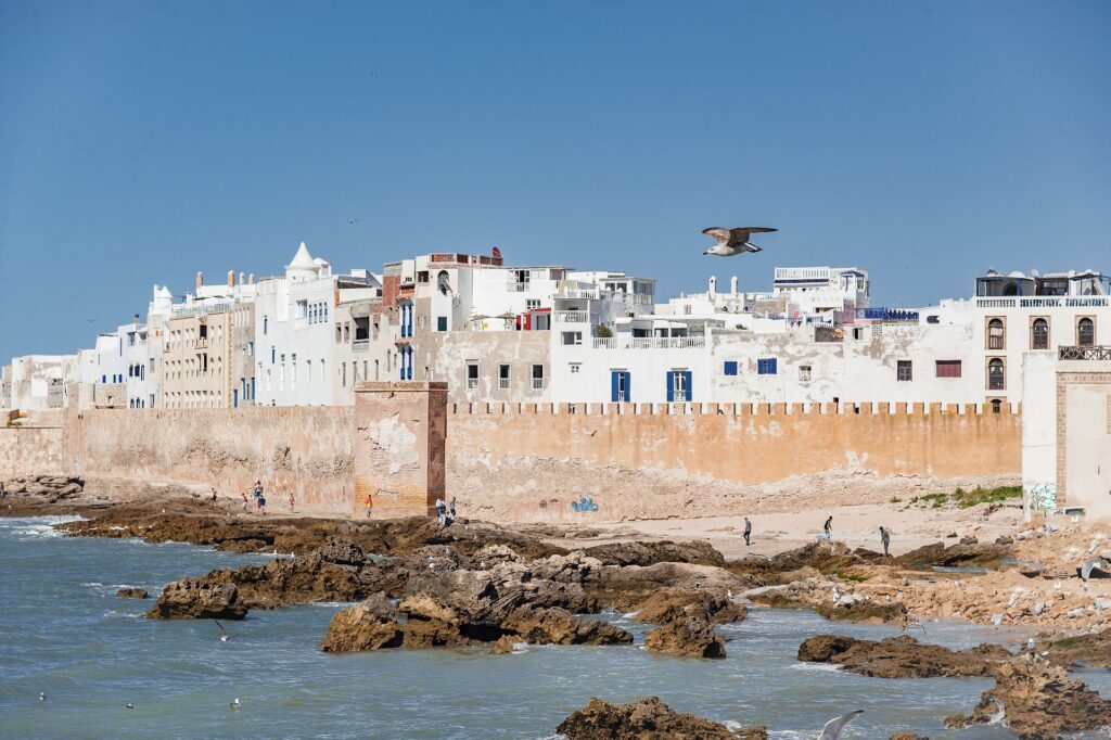 Vue des remparts d’Essaouira avec l’océan Atlantique en avant-plan et des personnes profitant de la plage.